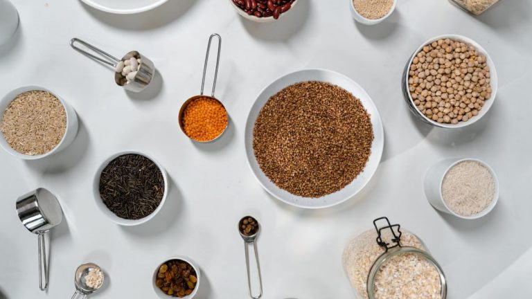 Top view of various grains, legumes, and spices in bowls and jars on a white table.